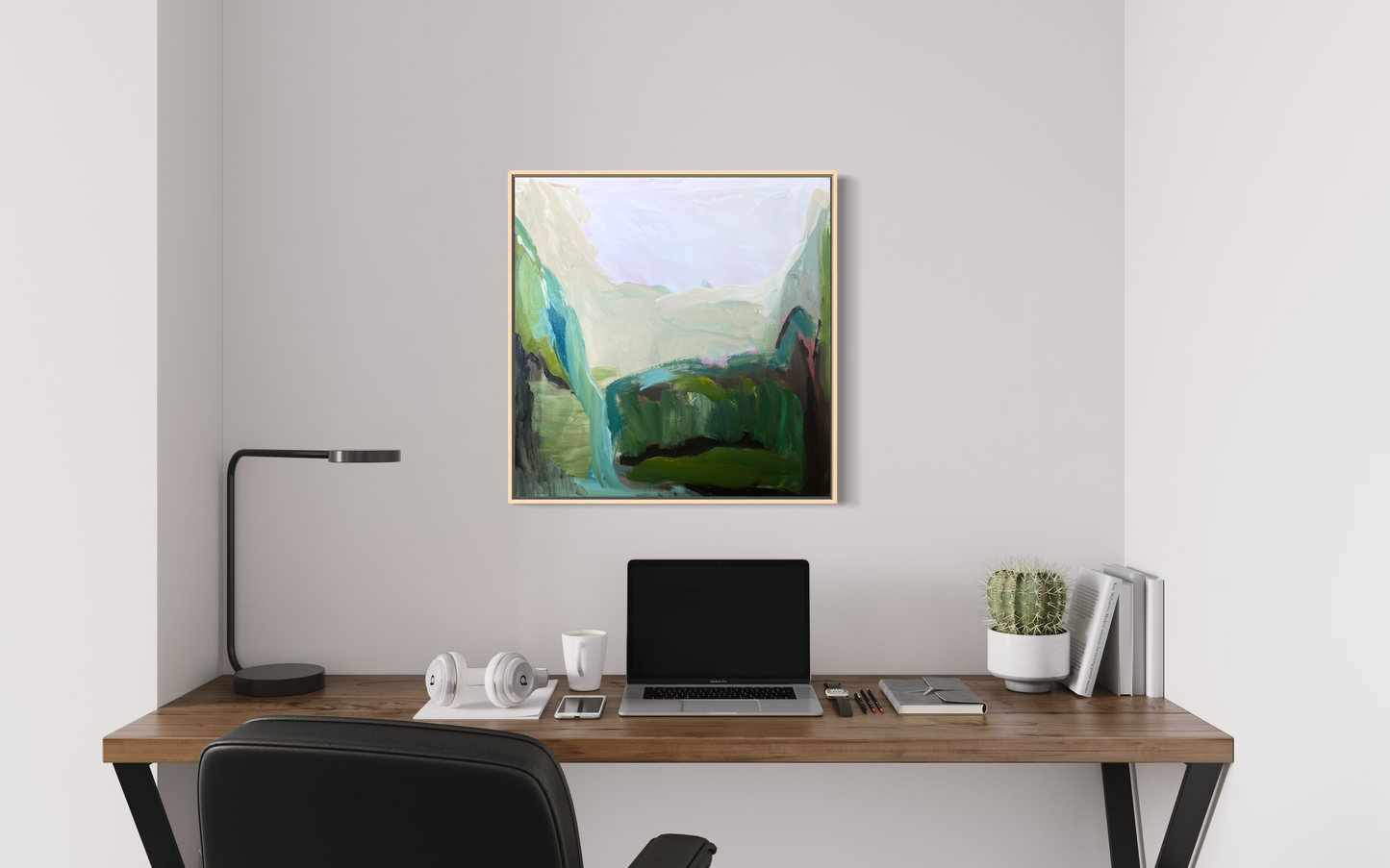 Modern office desk with a laptop, lamp, and decorative items against a white wall with a framed abstract landscape painting.